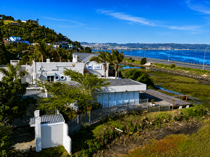 Aerial view of the property featuring a timber deck, metal fencing, and flat roof architecture, situated near the estuary and coastal road.