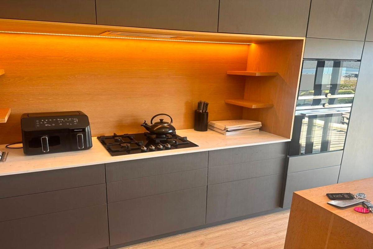 Modern kitchen featuring sleek dark cabinetry, stone countertops, integrated oven, gas cooktop, and warm under-cabinet lighting with floating timber shelving.