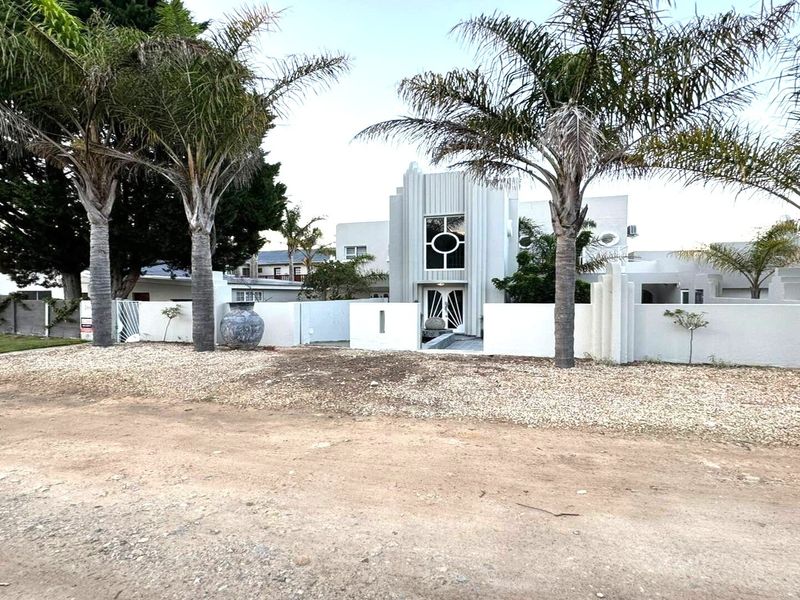 Front elevation of a modern architectural house featuring a distinctive central facade, gated entrance, and established palm trees set behind a boundary wall.