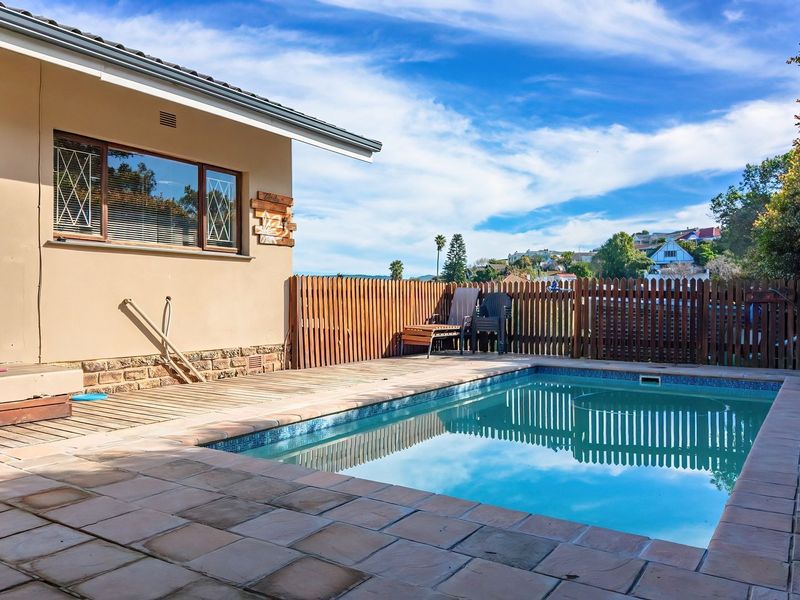 Outdoor pool area featuring a swimming pool, tiled surround, wooden decking, and a timber picket fence, set against a clear blue sky.