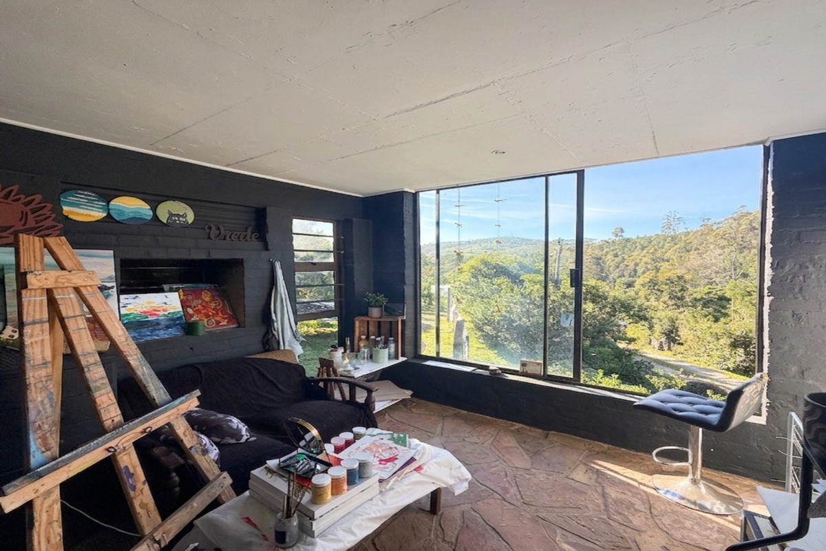A sunlit living space featuring rustic stone flooring, dark painted brick walls, and a large window offering expansive views of the surrounding landscape.