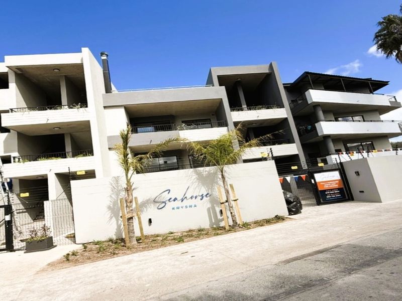 Modern apartment complex facade featuring multi-level balconies, secure boundary wall with electric fencing, and a paved entrance area.