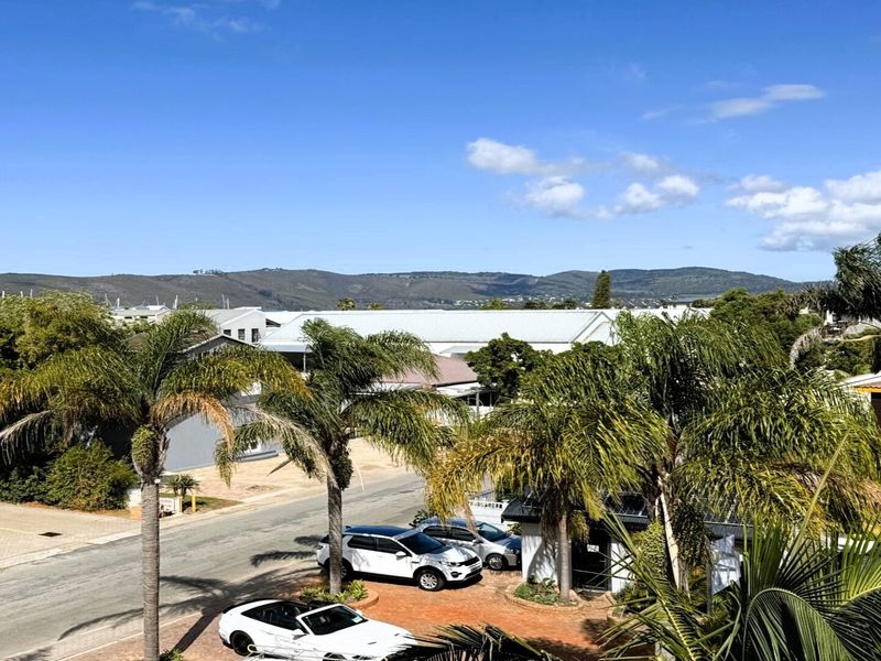 An elevated outdoor view overlooking a suburban street, surrounding trees, and distant mountain ranges under a clear blue sky.