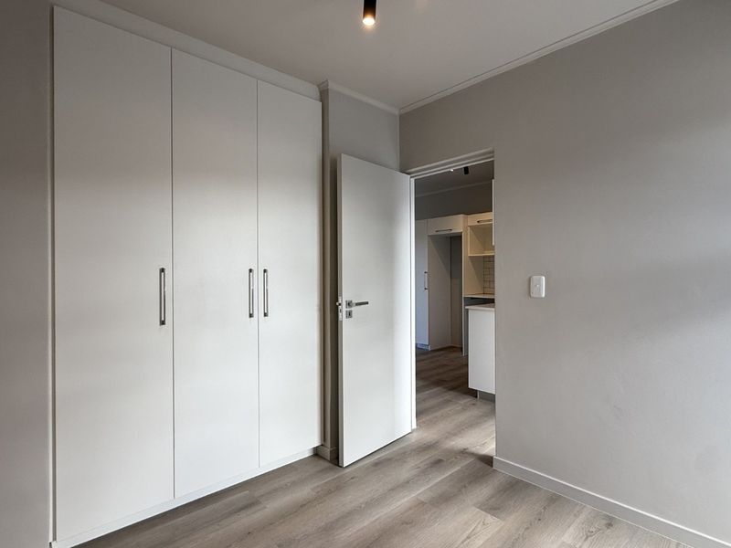 A clean, modern bedroom featuring neutral walls, light laminate flooring, and ample built-in cupboard storage, with a view into the adjacent kitchen area.