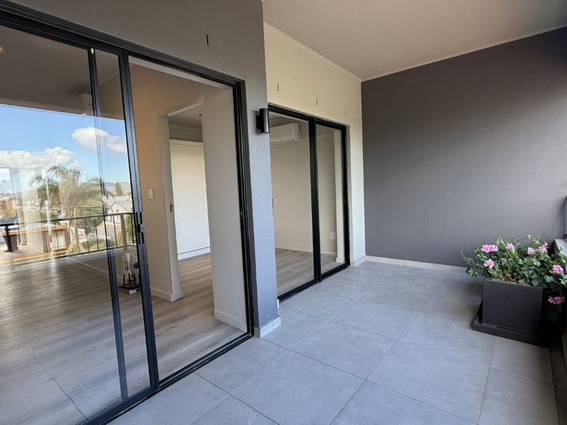 A modern, covered balcony featuring large sliding glass doors, neutral tiled flooring, and a sleek metal balustrade with views of the surrounding area.