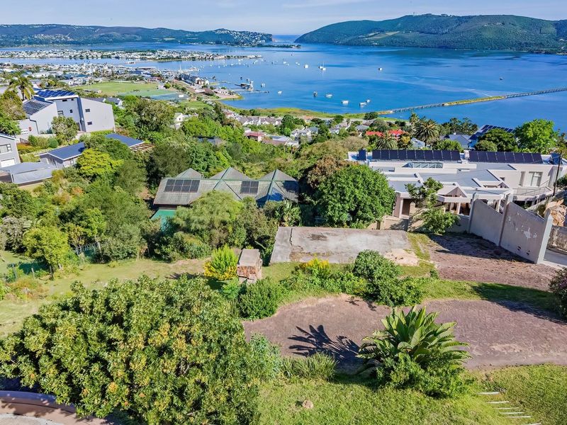 Aerial view of a vacant residential plot featuring a concrete slab foundation and retaining wall, overlooking the scenic Knysna lagoon and surrounding neighborhood.