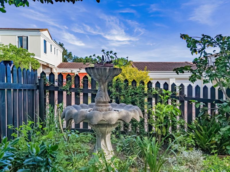 A well-maintained garden area featuring a decorative stone water fountain and a dark timber picket fence with residential buildings in the background.