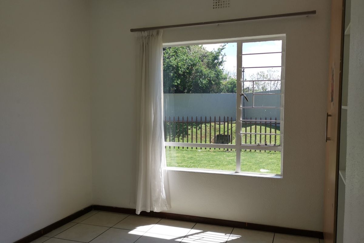 A bright, empty bedroom featuring tiled flooring, a built-in cupboard, and a large window with security bars overlooking the garden.