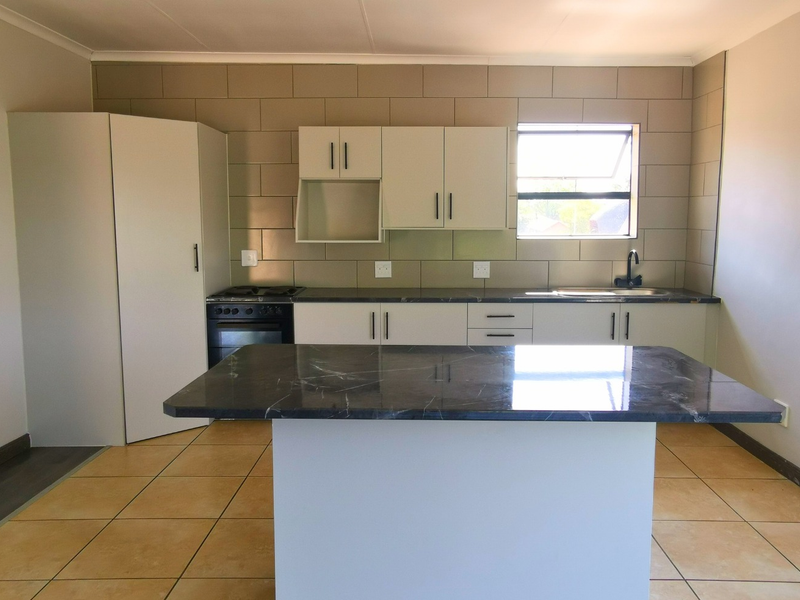 A clean, modern kitchen featuring a central island, built-in cabinetry, stone countertops, and integrated appliances, finished with neutral tiling.