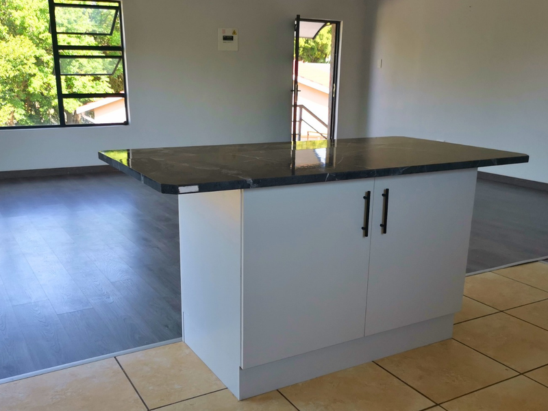 An open-plan living area featuring a central kitchen island with a dark stone countertop, contrasting laminate and ceramic tile flooring, and large casement windows.