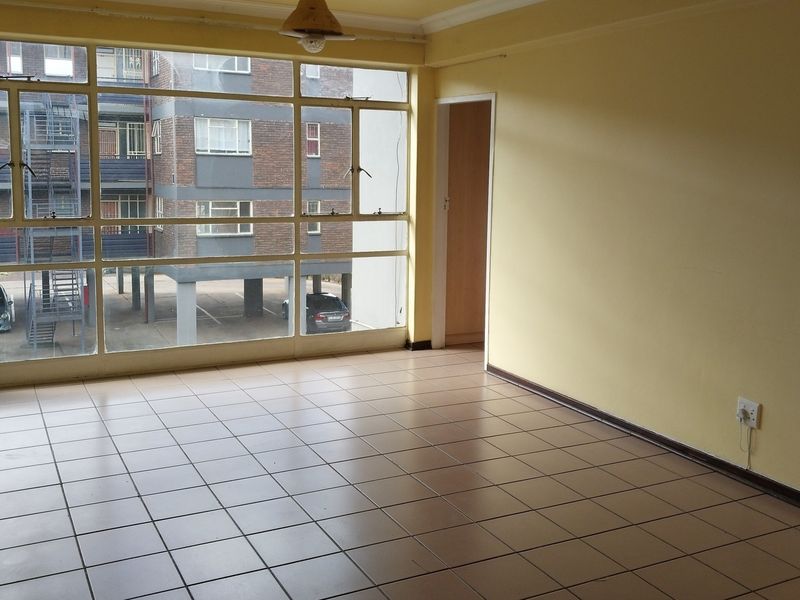 An empty living room featuring tiled flooring and large steel-framed windows that provide natural light and a view of the surrounding apartment complex.