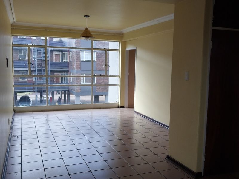An empty, sunlit living area featuring neutral tiled flooring, coved ceilings, and large windows providing ample natural light.