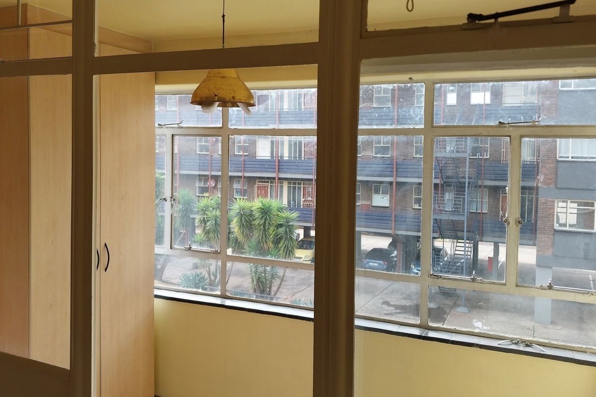 A vacant bedroom featuring built-in cupboards and large steel-framed windows overlooking the complex courtyard.