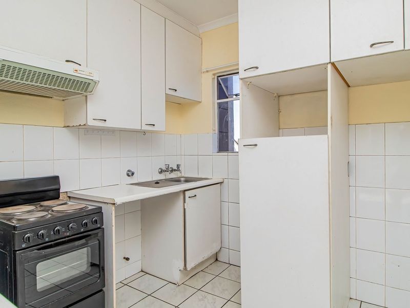 A compact, dated kitchen featuring white cabinetry, tiled splashbacks, a stainless steel sink, and a freestanding stove, offering potential for modernization.