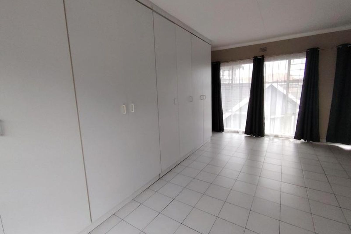 A spacious, empty bedroom featuring extensive built-in cupboards, neutral tiled flooring, and large windows with security bars.