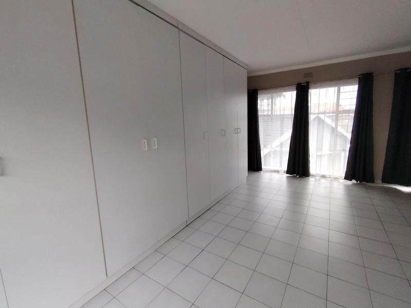 A spacious, empty bedroom featuring extensive built-in cupboards, neutral tiled flooring, and large windows with security bars.
