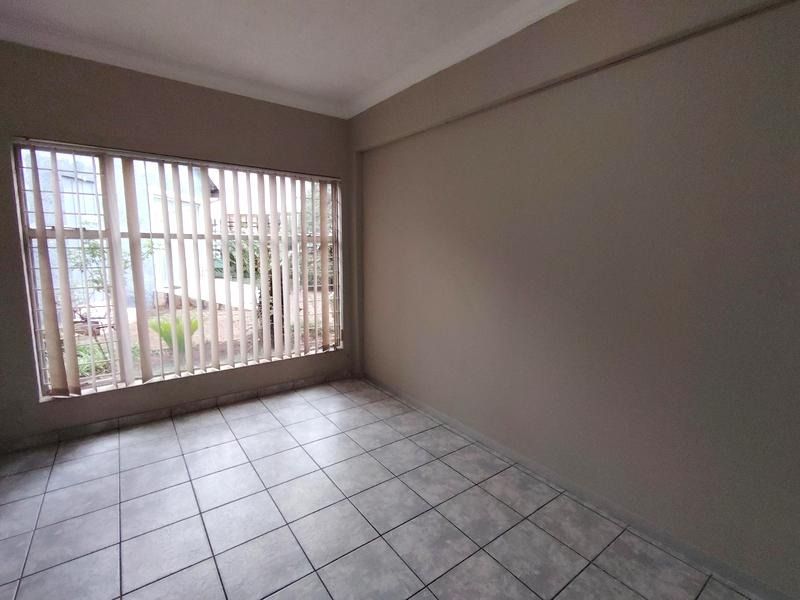A bright, empty bedroom featuring neutral wall tones, tiled flooring, and large windows fitted with vertical blinds.
