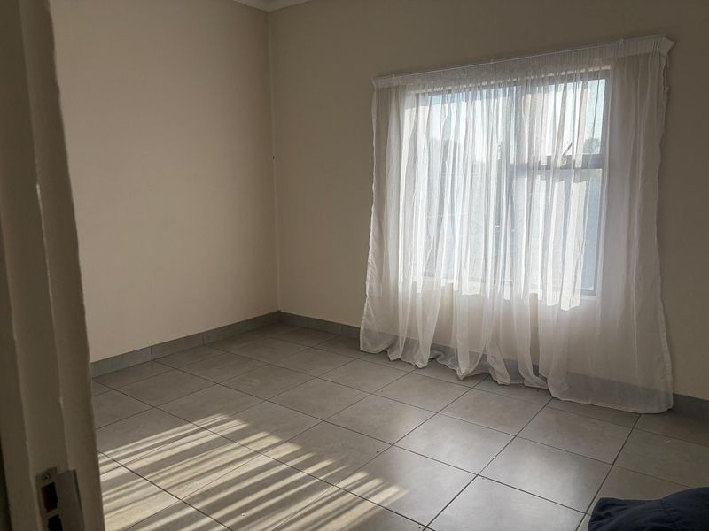 An empty bedroom featuring neutral tiled flooring and a window fitted with a curtain rail and sheer curtains.