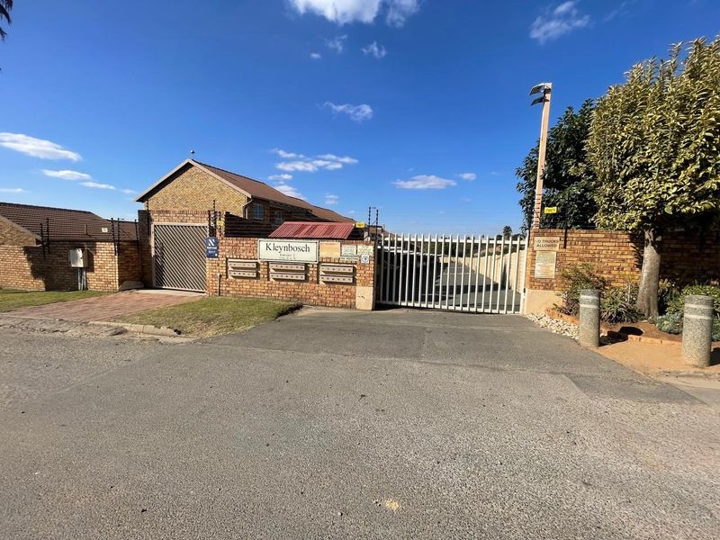 Front entrance view of Kleynbosch apartment complex featuring a secure automated gate, brick boundary wall, electric fencing, and communal post boxes.