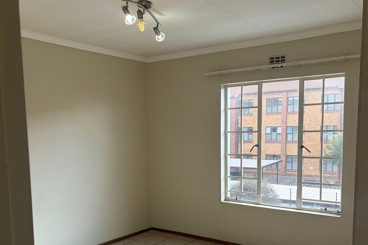 A bright, empty bedroom featuring tiled flooring, a large steel-framed window with a curtain rail, and a modern ceiling light fixture.