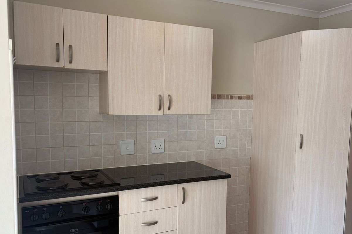 A clean, functional kitchen featuring light wood-finish cabinetry, dark laminate countertops, a built-in electric oven and hob, and a tiled backsplash.