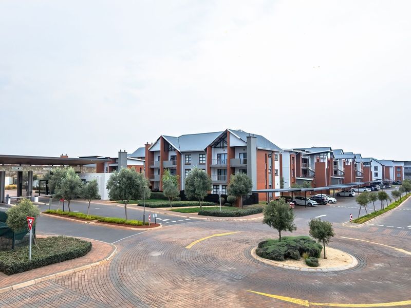 Wide-angle view of the apartment complex entrance, featuring paved driveways, landscaped gardens, security gatehouse, and multiple residential apartment blocks with covered parking.