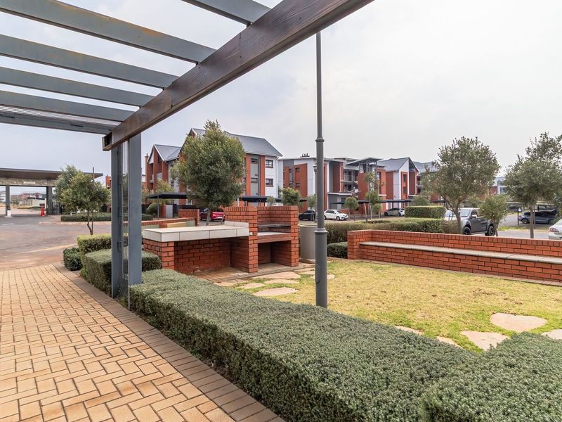 Communal outdoor area featuring a brick built-in braai, brick bench seating, and a pergola, set within a well-maintained residential apartment complex.