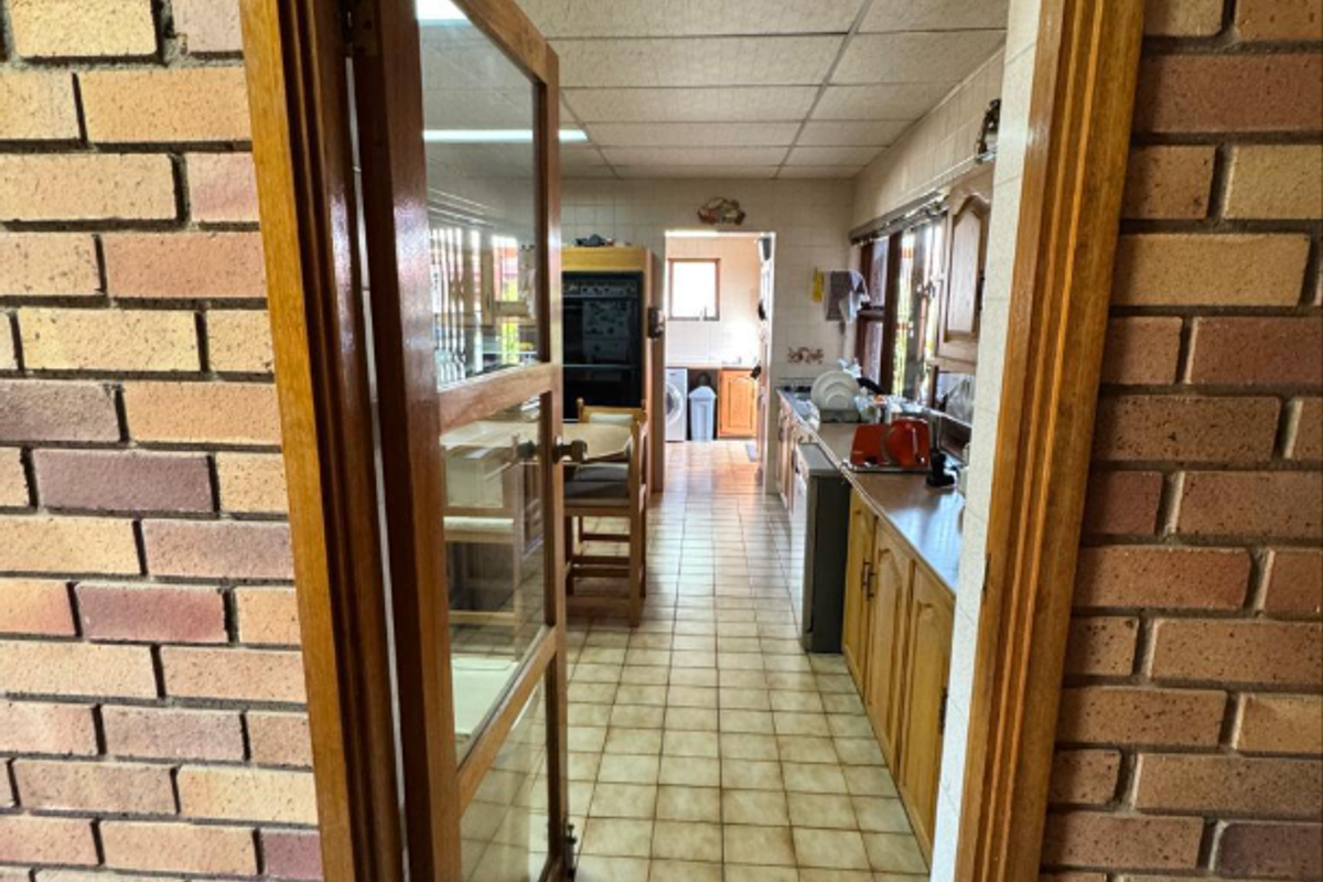A view through a timber doorway into a dated kitchen featuring tiled flooring, built-in cabinetry, and a drop ceiling.