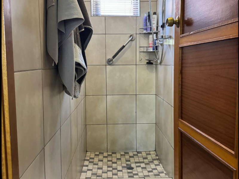 A compact shower enclosure featuring neutral wall tiling, a mosaic-tiled shower floor, a fitted shower head, and a safety grab rail.