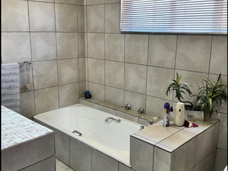 A functional bathroom featuring a built-in tiled bathtub, neutral wall and floor tiling, and a window with fitted blinds.