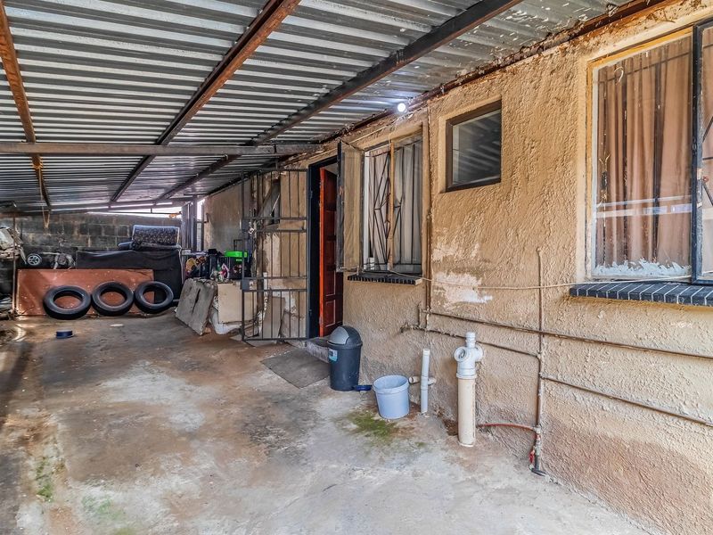A covered outdoor patio area featuring a corrugated metal roof, security bars on windows, and concrete flooring, requiring maintenance and cosmetic updates.