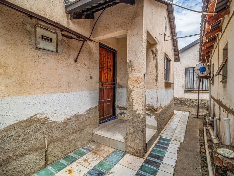 A narrow exterior side passage featuring a tiled walkway, security gate, and visible exterior plumbing, showing significant wear to the wall plaster and finishes.