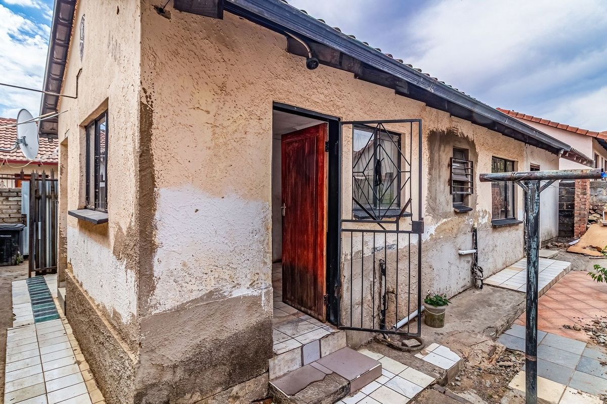 Rear exterior view of the property showing a weathered facade with peeling plaster, security gate, and tiled patio area requiring maintenance.