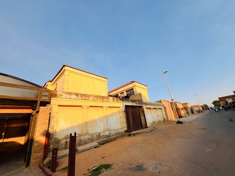 Street view of the property featuring a boundary wall with decorative columns and a metal gate, showing signs of exterior wear and weathering.