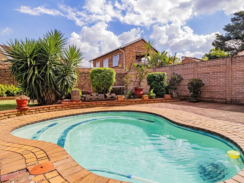 A well-maintained outdoor pool area featuring a curved swimming pool, paved surround, and raised brick planter beds enclosed by a secure brick boundary wall.