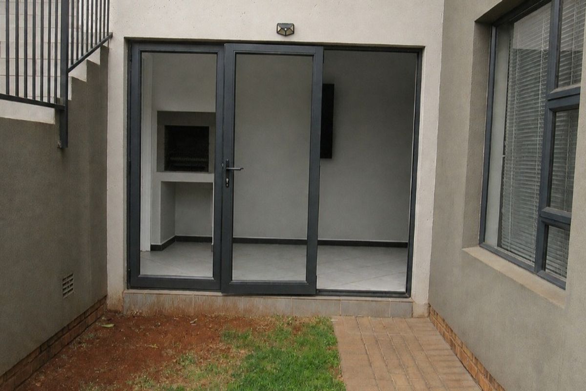 Rear exterior view of the townhouse featuring a paved walkway, small lawn area, and access to an indoor braai area through aluminium sliding doors.