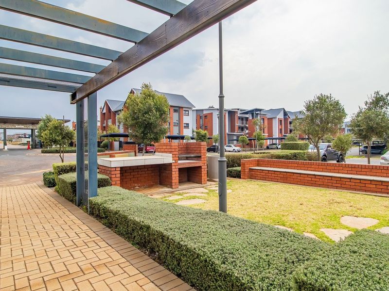 A well-maintained communal garden area featuring a brick built-in braai, paved walkways, and a pergola within a residential townhouse complex.
