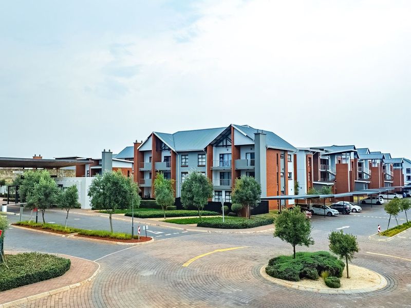 A wide-angle view of a modern townhouse complex entrance, featuring paved driveways, landscaped gardens, and covered parking structures.