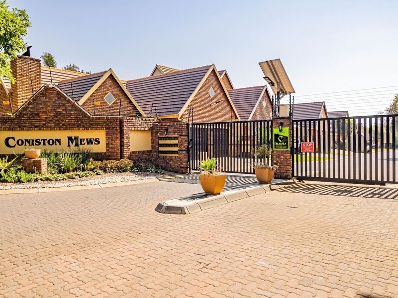 The secure entrance to Coniston Mews townhouse complex, featuring a brick facade, automated sliding gate, electric fencing, and a well-maintained paved driveway.