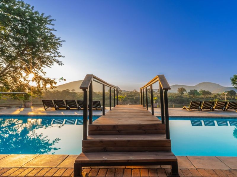 A scenic outdoor pool area featuring a wooden bridge walkway, paved decking, and expansive views of the surrounding landscape and mountains.