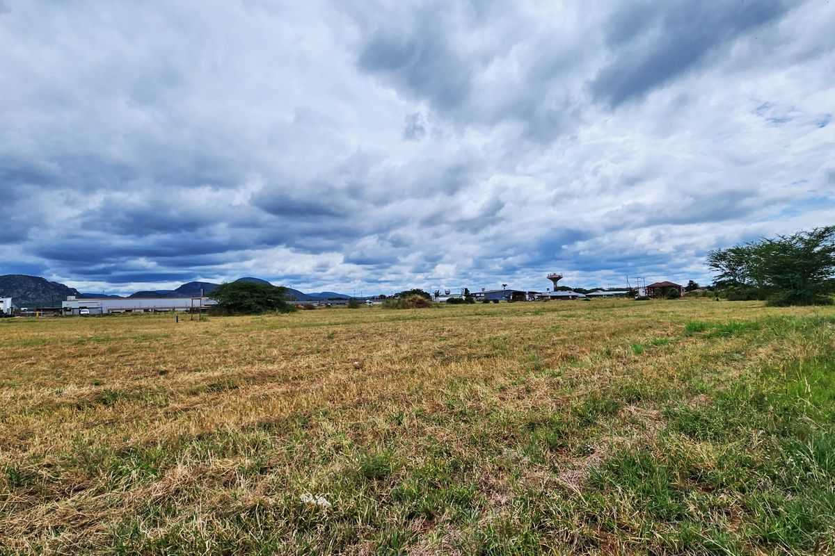 A wide-angle view of a vacant commercial land plot featuring open grassland with distant industrial structures and mountain views under a cloudy sky.