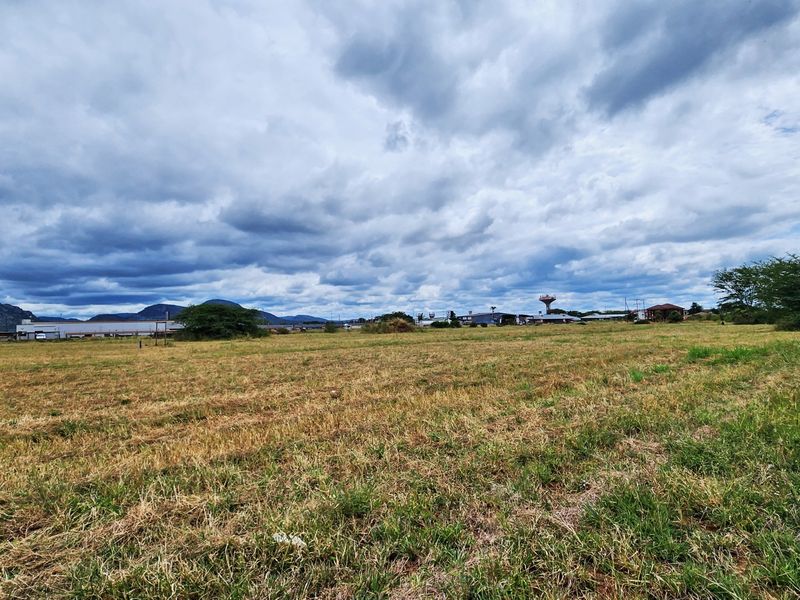 A wide-angle view of a vacant commercial land plot featuring open grassland with distant industrial structures and mountain views under a cloudy sky.