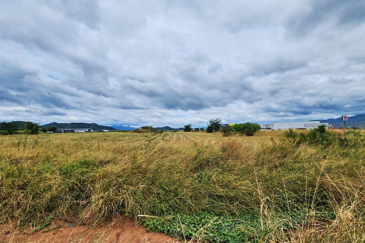 A wide-angle view of a vacant commercial land plot featuring open grassland with distant mountain views under a cloudy sky.