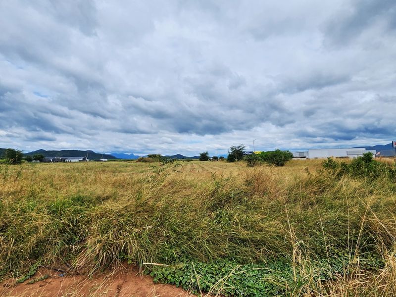 A wide-angle view of a vacant commercial land plot featuring open grassland with distant mountain views under a cloudy sky.