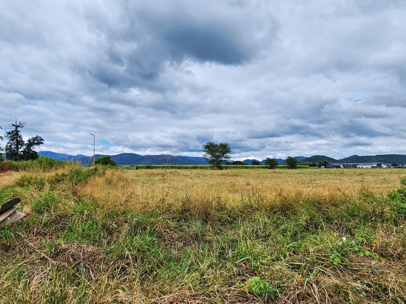 A wide-angle view of a vacant commercial land plot featuring natural grassland, perimeter fencing, and distant mountain views.