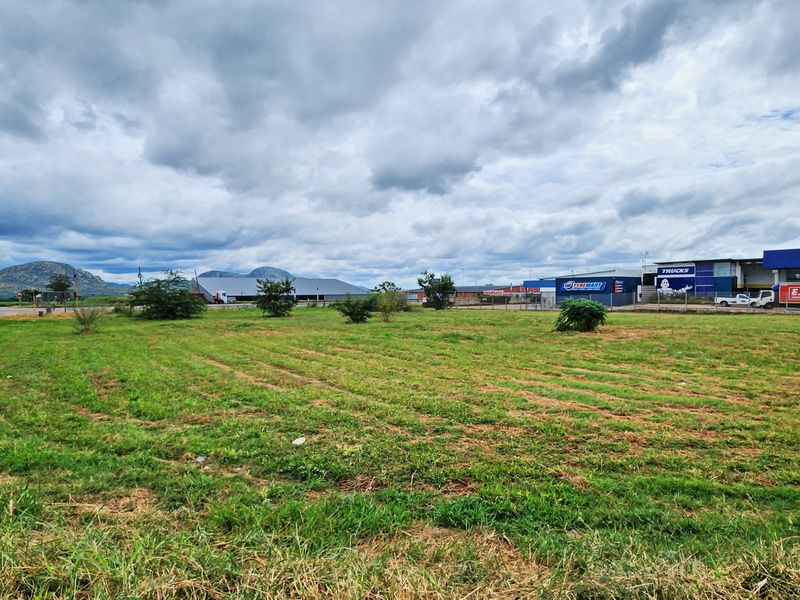 A vacant commercial land plot featuring open grass terrain, situated adjacent to established commercial and industrial service centers.