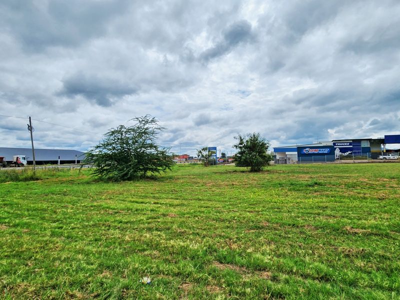 A wide-angle view of a vacant commercial land plot featuring open grass areas with surrounding commercial infrastructure and utility poles.