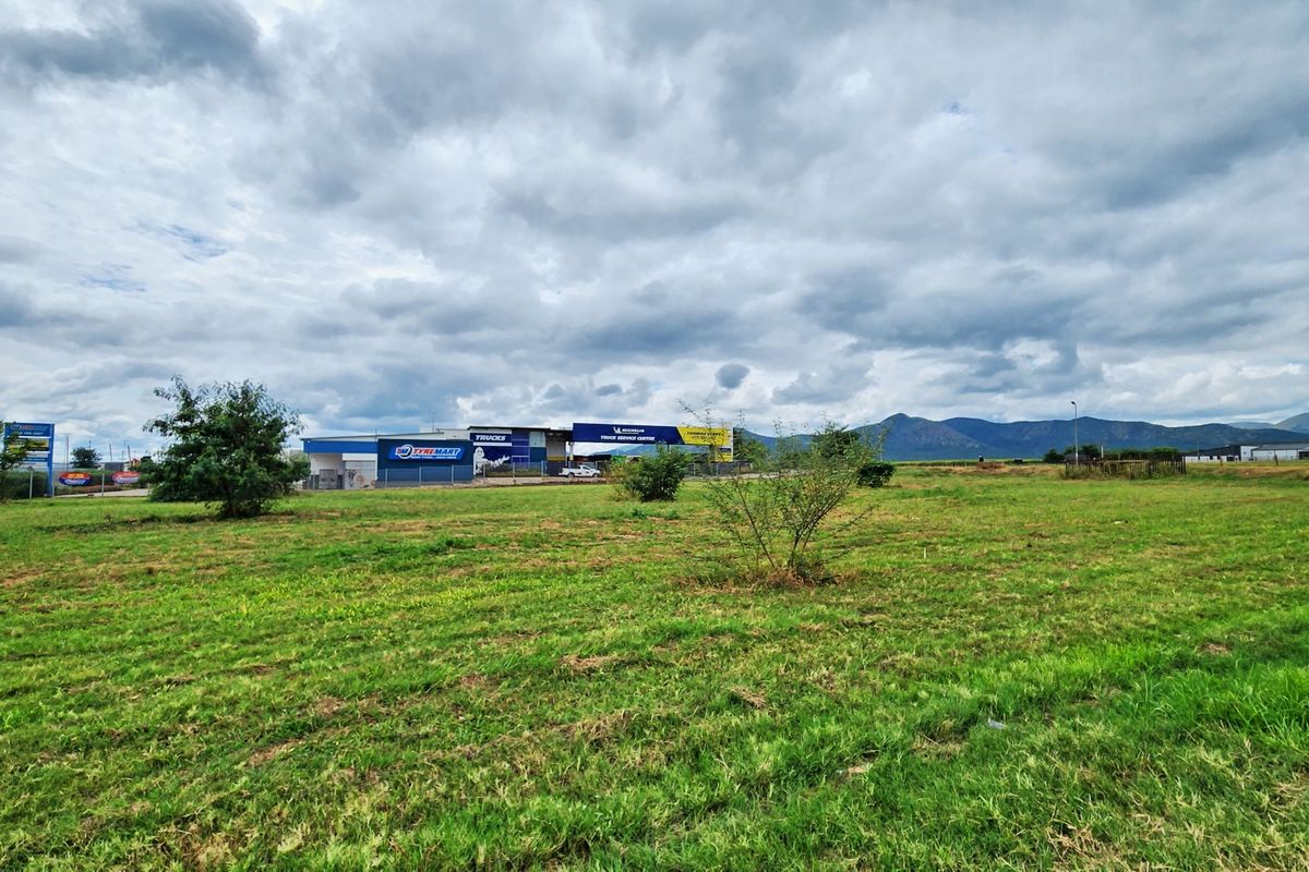A wide-angle view of a vacant commercial land plot featuring open grass fields with surrounding commercial infrastructure and mountain views in the distance.