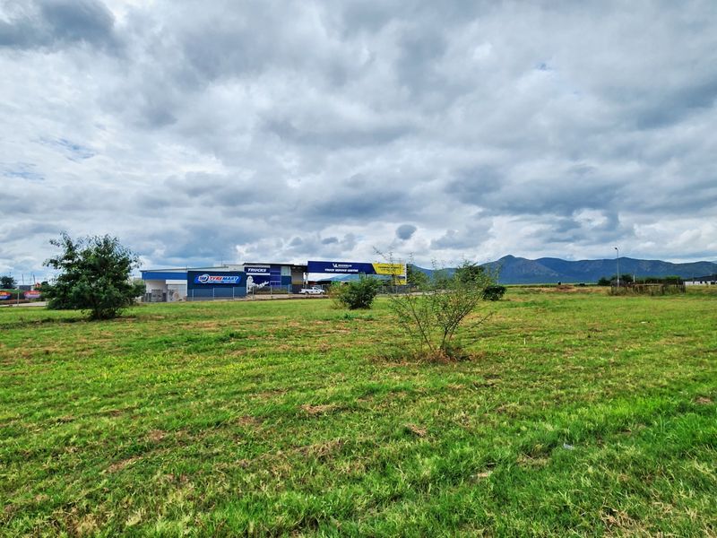A wide-angle view of a vacant commercial land plot featuring open grass fields with surrounding commercial infrastructure and mountain views in the distance.