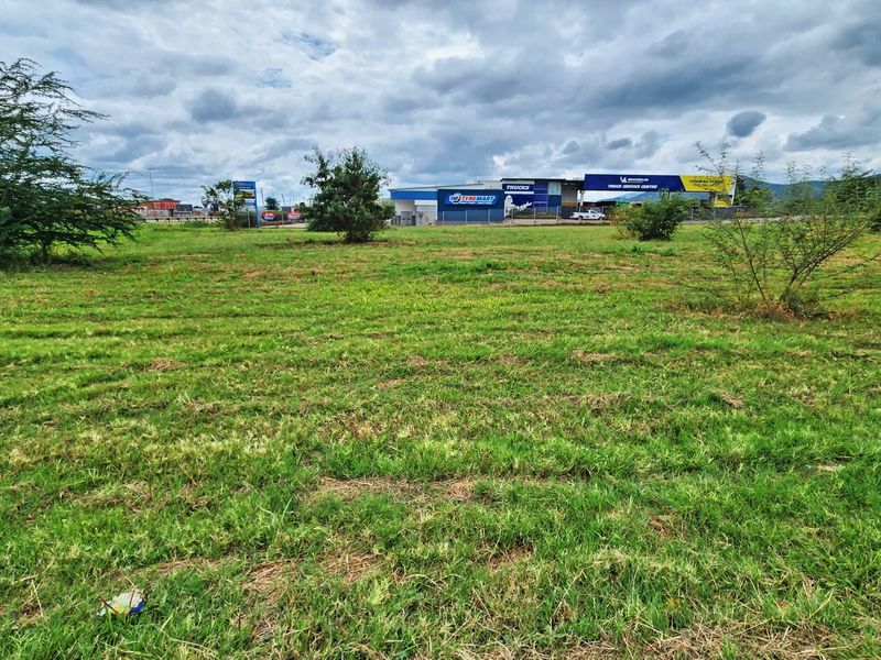 A wide-angle view of a vacant commercial land plot with established grass and nearby commercial infrastructure in the background.